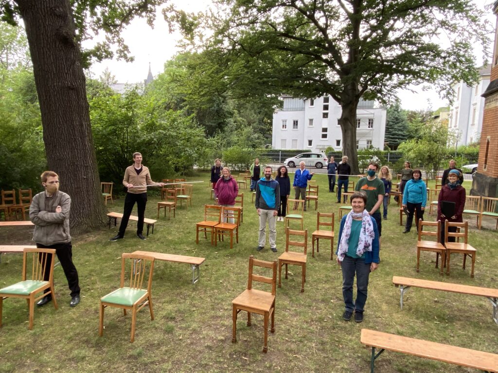 Chorsänger stehen mit großem Abstand und zum Teil mit Masken im Freien zwischen einzelnen Stühlen. Ein Chorsänger hält einen Zollstock als Abstandsmaß.