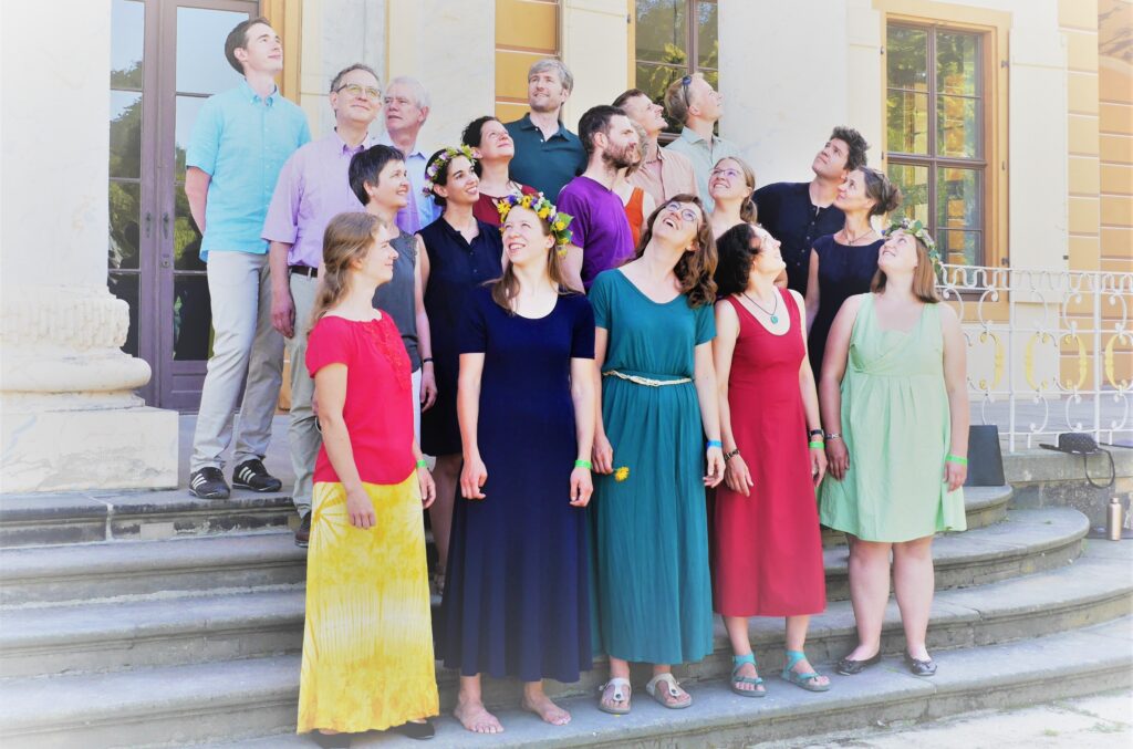 19 Sängerinnen und Sänger des Kammerchors anima nordica stehen auf einer Außentreppe, lächeln und schauen in verschiedene Richtungen in den Himmel. Sie tragen bunte Sommerkleider und Freizeitkleidung.
